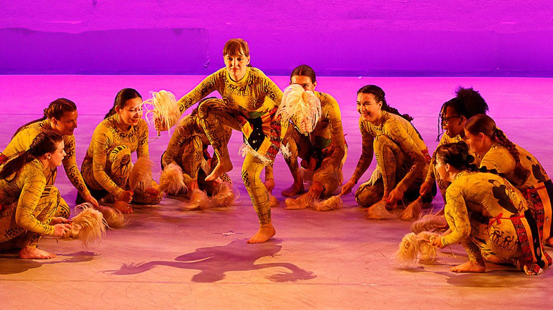 Several dancers in brightly colored costumes sit on the floor to encircle a soloist standing with a lifted, flexed foot and bunches of feathers in each hand. The dancers perform a representation of the West African dance Zaouli in There is no movement without rhythm, choreographed by Webster Dance faculty member Diadie Bathily.