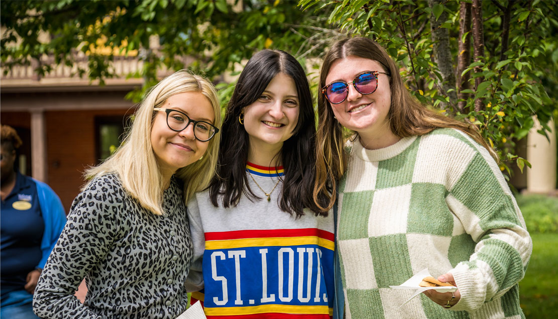 Three young women stand and smile together at Webster event, one wears sweater that reads "St. Louis."