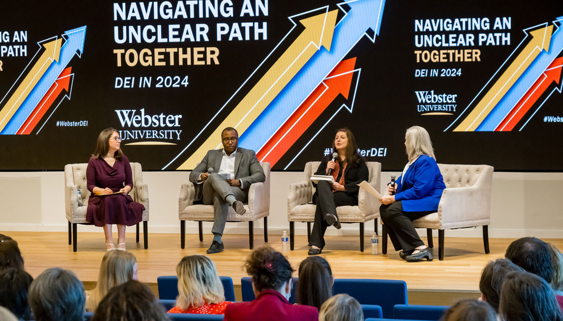 Panelists dressed in business attire are sitting in armchairs on a stage.  The screen behind the panelists has the title "Navigating an Unclear Path Together" and the logo of the University.