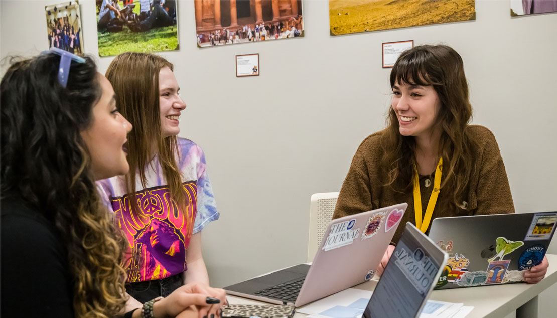 Three students sit at a table with laptops, working together.