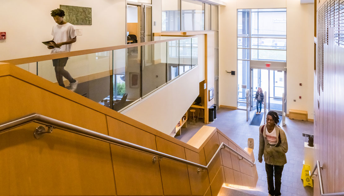 View from top of staircase of student walking up with books and another walking on top floor.