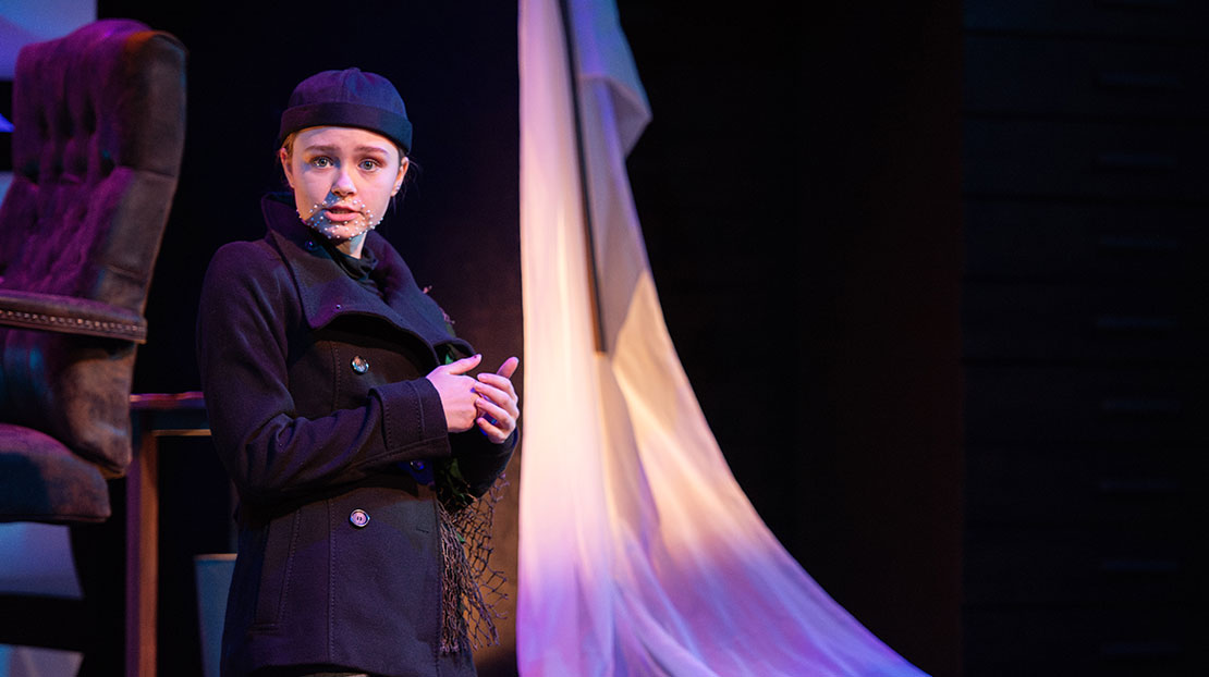 An actor wearing a peacoat and stocking cap, has a beard made of pearls and a sleeve with fishing net. Behind the actor is a chair and a hanging draped cloth.