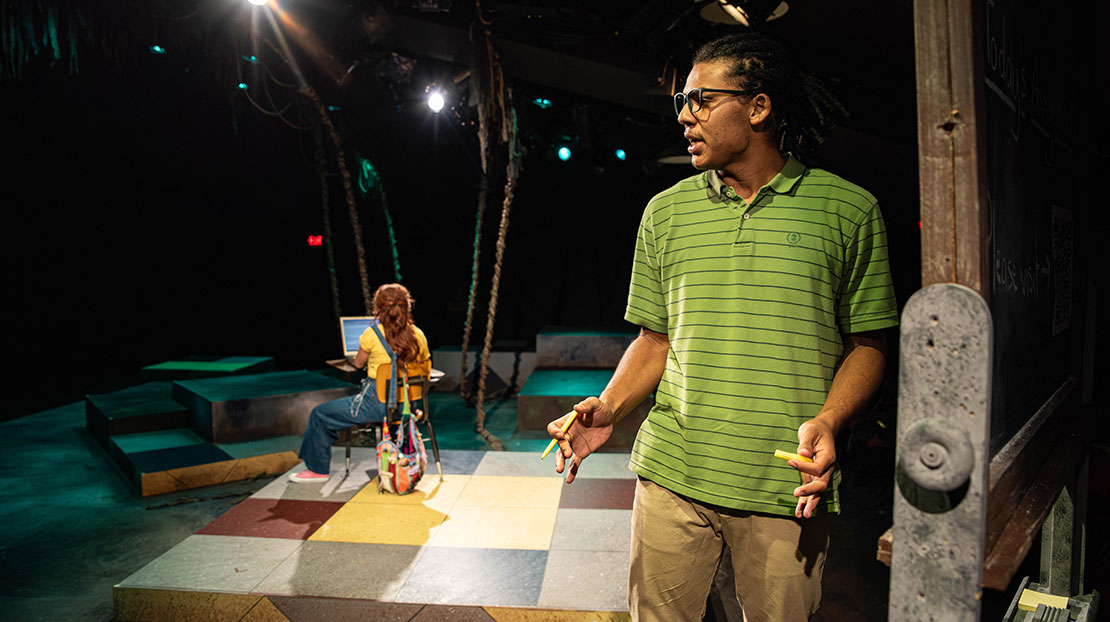 A multi-level stage with two actors: a man performs a serious monologue while a woman sitting in a chair has her back to him. She is looking at a computer.