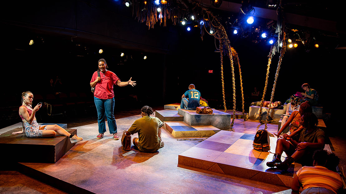 Actors sit on small, multi-level stages painted in earth-toned, grid work. One actor stands with a microphone in her hand.