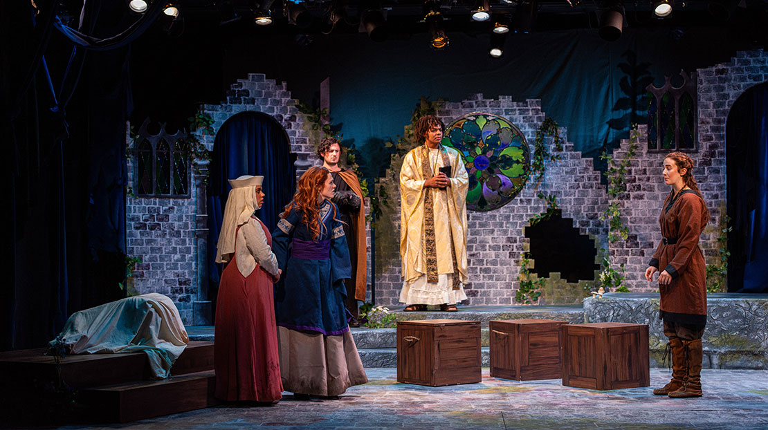 Five actors dressed in medieval clothing stand in front of a crumbling stone wall and large, round, stained-glass window.