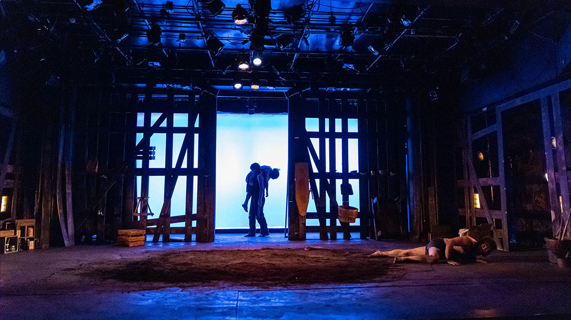 An actor lays on a dirt floor. In the back, another actor carries someone over their shoulder. They stand in front of a blue-lit screen with a wooden framework structure on both sides.