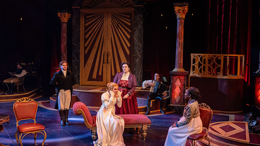 A woman in a period dress sits on a red velvet chaise lounge surrounded by other actors and an accordion player. 