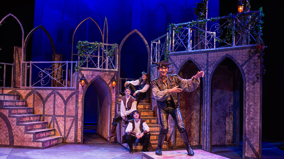 An actor with horns wearing black leather stands in front of three others who sit on stairs behind him. The set has archways and stairs leading to a balcony with decorative, metalwork railing and climbing flowered vines.