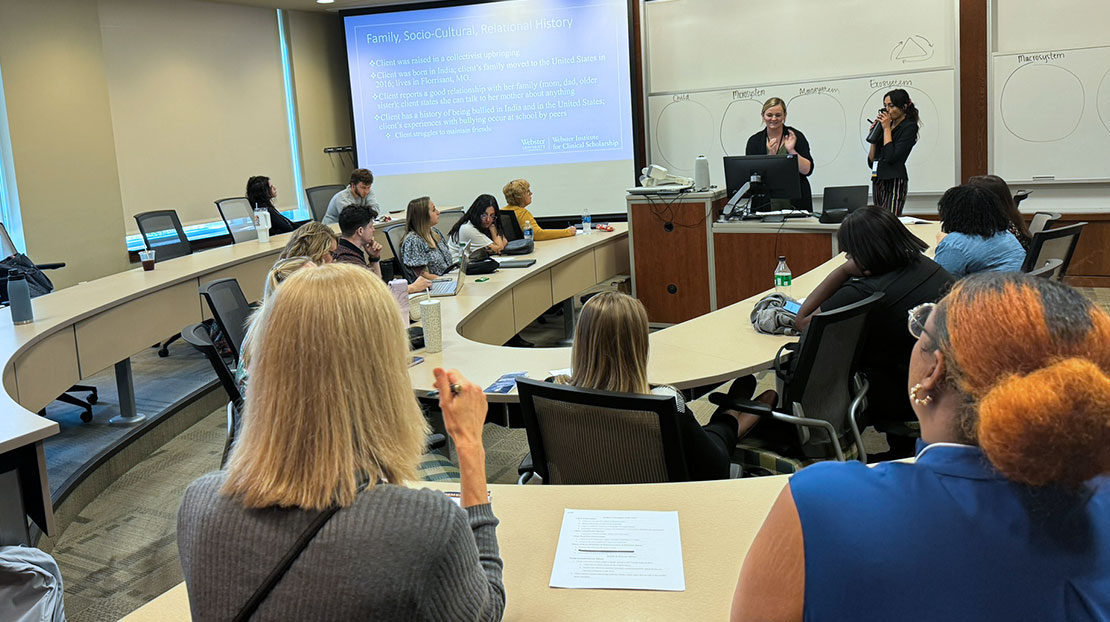 Presenters stand in front of classroom with attendees sitting at curved tables.