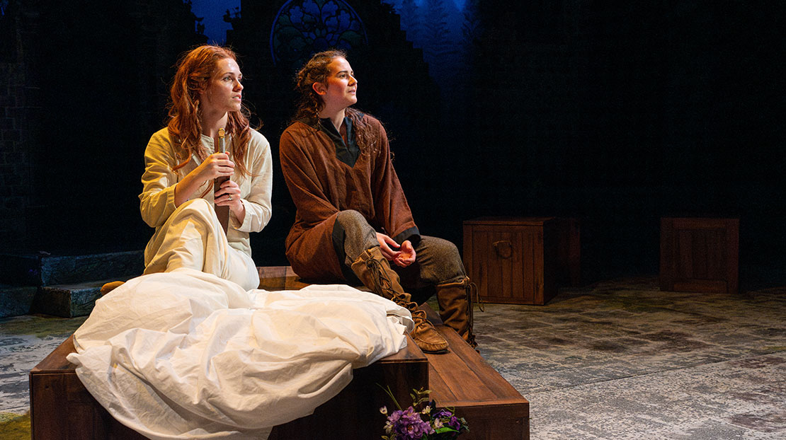 Two actors sit next to each other on stage looking in the distance. One on the left wears a white gown and holds dagger, the other wears brown tunic and boots.