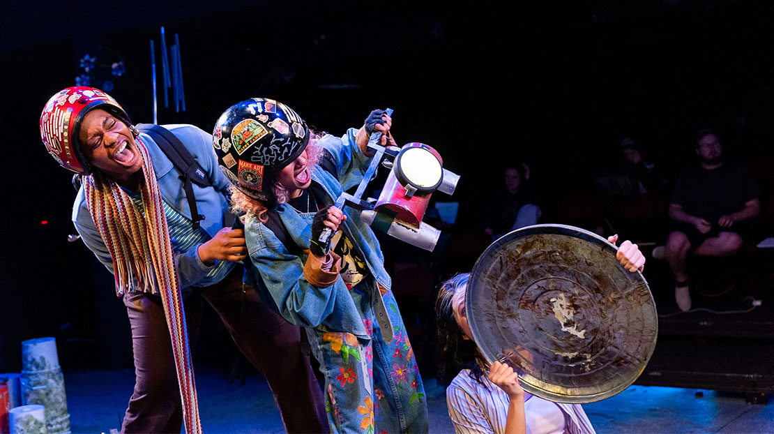 Three actors are onstage. Two wear sticker-laden helmets and mimic driving. One actor holds a trash can lid, while the other holds an old bike handle. 