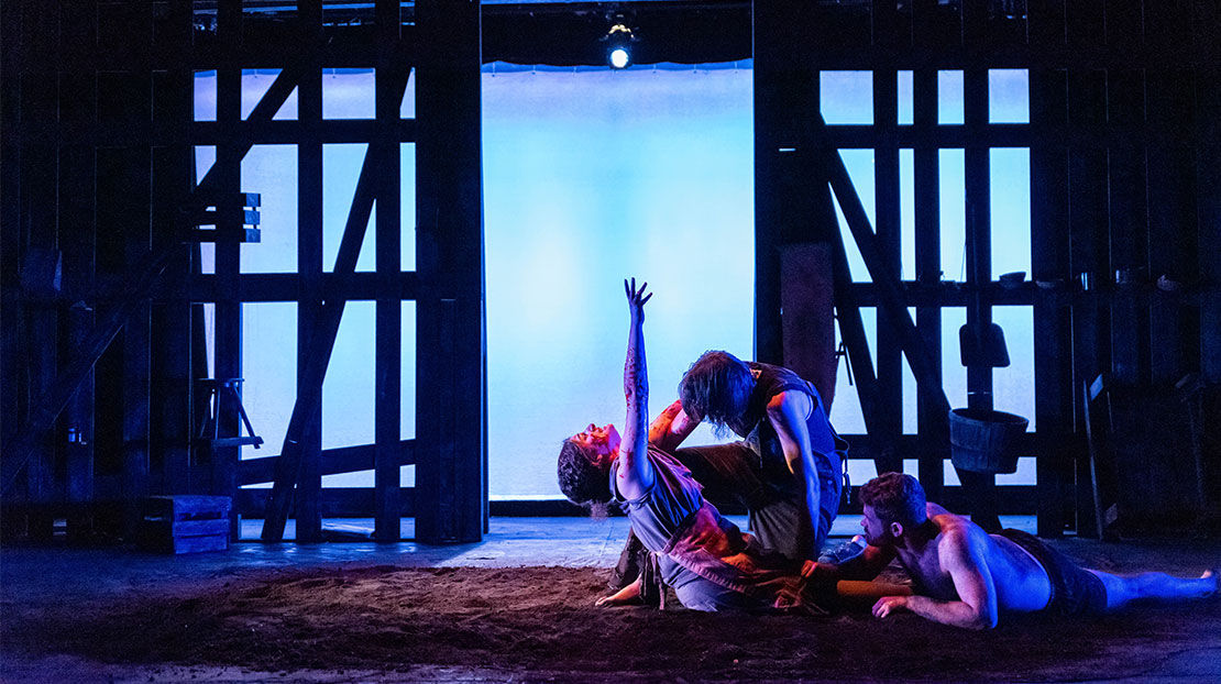 Three actors in dramatic poses sit, lay, and kneel together on a dirt floor. The background is a blue-lit screen with a wooden framework structure in front.