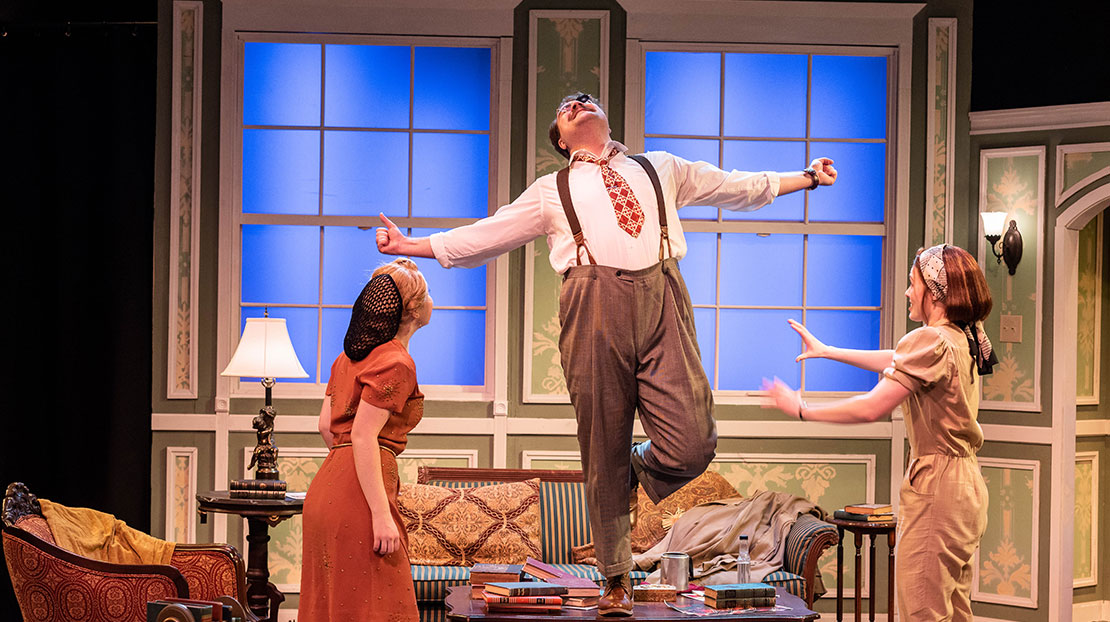 In a living room, a man in a suit and suspenders stands on one leg on top of a coffee table with his arms out to the side and head back while two women on the floor in front appear worried he will fall.
