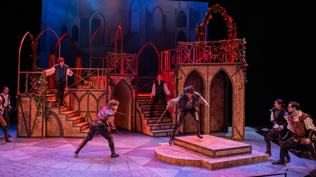 Two actors are in the middle of a stage engaged in a swordfight, while other actors, also with swords, stand on multi-level staircases in the background. 