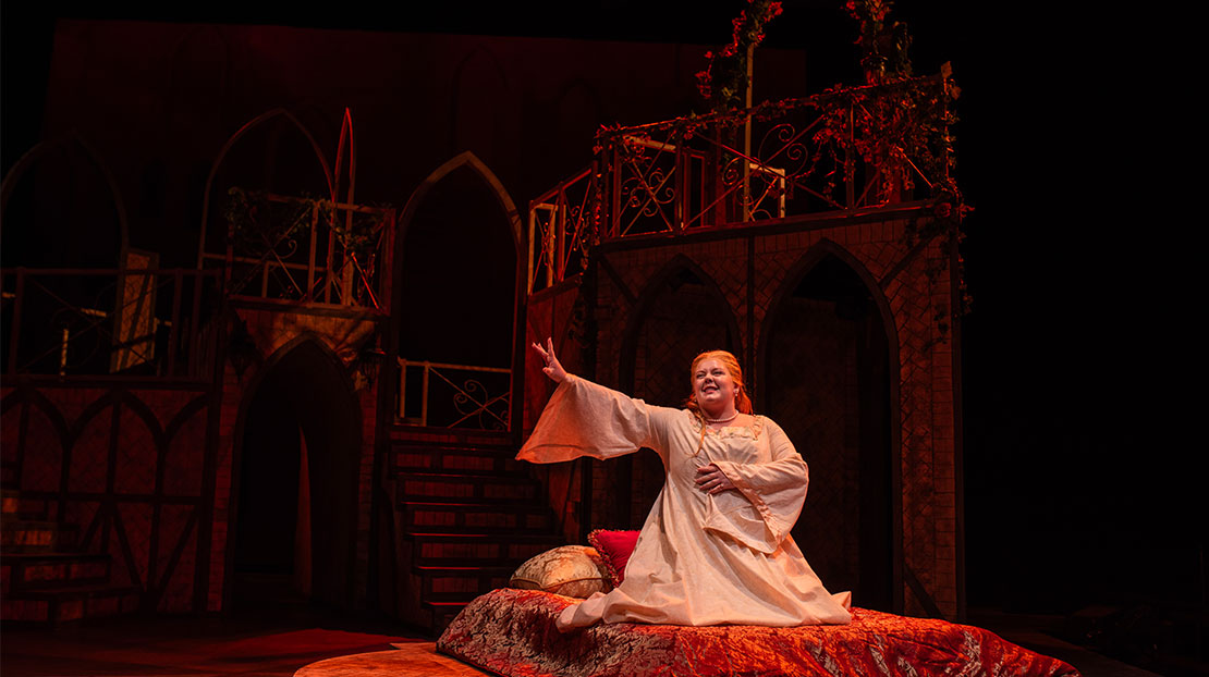 A woman dressed in a white nightgown kneels on a red velvet bed with gold brocade pillows in the center of the stage. In the background are dimly lit stairways and arches.