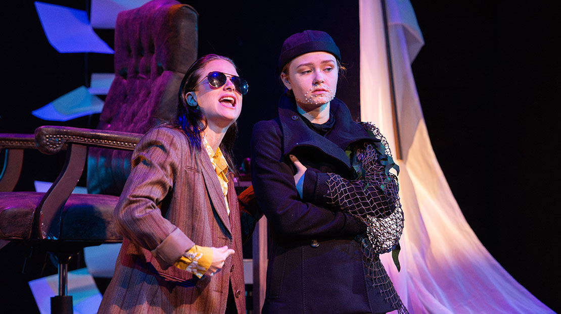 Two actors are dressed in Avant Garde, modern costumes. One actor wears sunglasses and the other has a beard made of pearls. Behind them, there is a leather chair and floating papers from floor to ceiling.