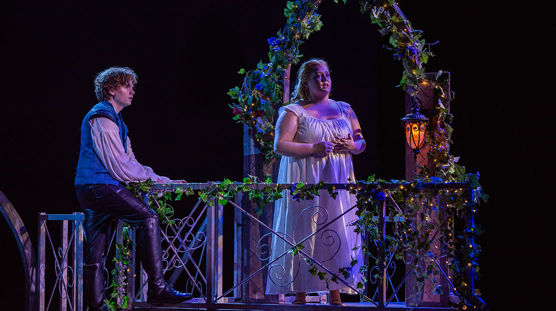 A woman stands on a flower-lined balcony with an arch in a long, flowing gown looks off to the distance, while a man stands to the side facing her. The stage lighting produces the effect of night through the interplay of light and shadow.
