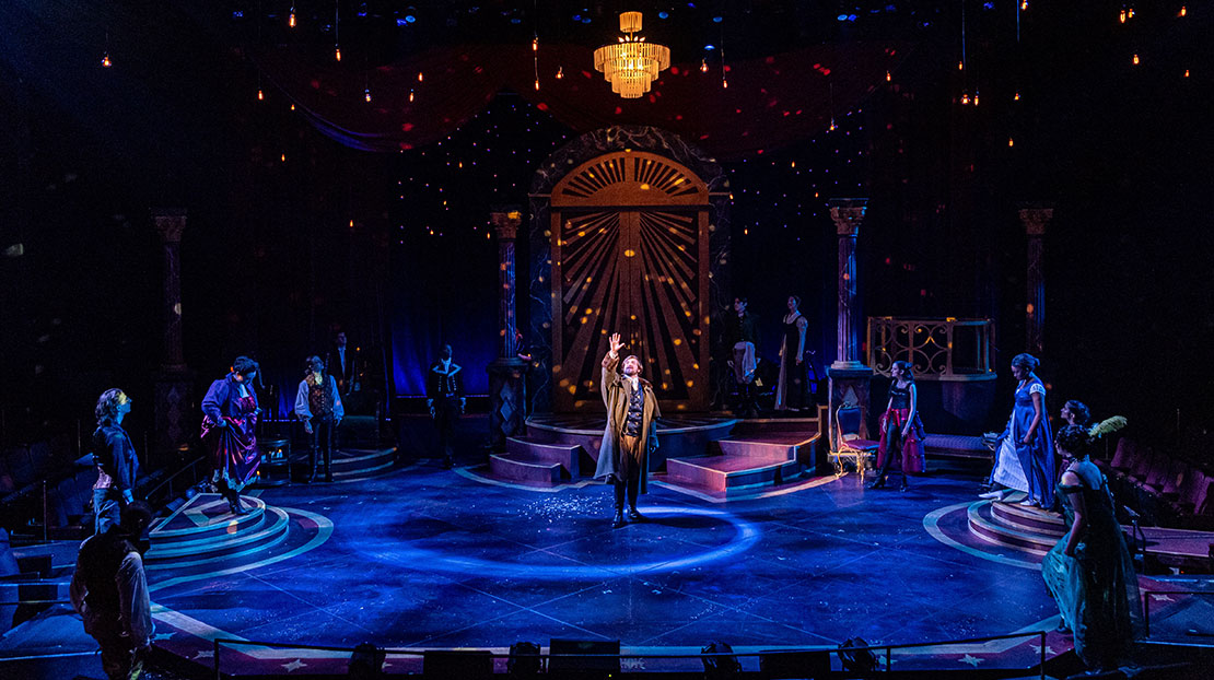 The stage lighting is dark blue with small, floating, multicolored red and yellow lights. An actor is in the center stage illuminated by a large glass chandelier and surrounded by a group of actors in the blue shadowed background.