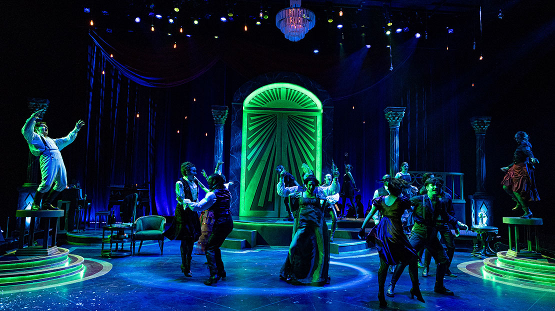 A large group of actors are having a raucous party, dancing on the floor and on tables in a furnished room. The stage lighting is dark blue with neon-dramatic green accents, in particular a huge neon door at the back of the stage.