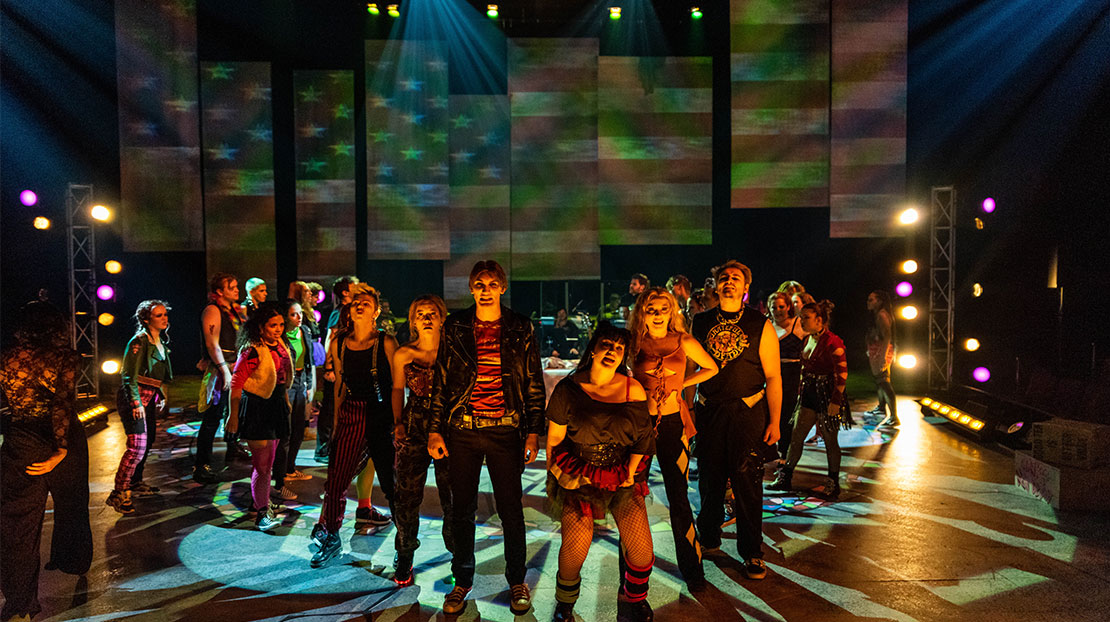 A large ensemble of actors on stage, dressed in punk clothing. The lighting is atmospheric and there are multiple beam angle lights which produce patterned shadows on the floor. A large American flag is projected onto a multi-paneled background.
