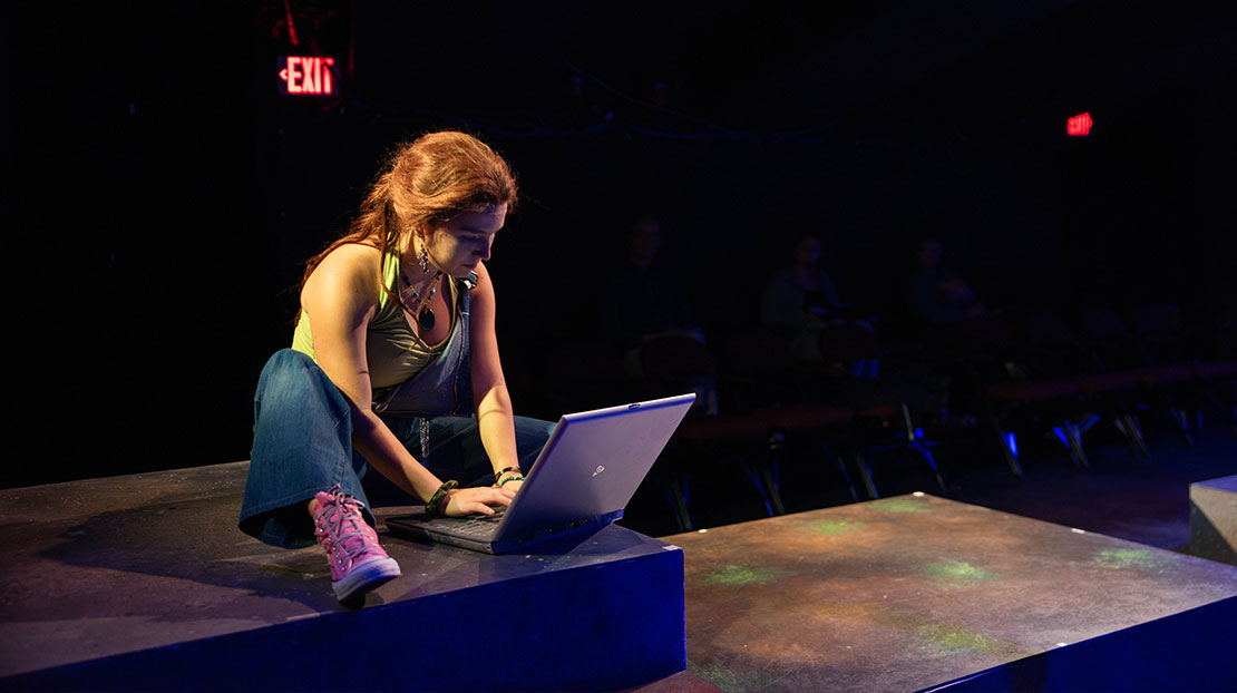 A modern-day woman sits on the floor and types on a laptop. The stage and background are dark, and the actor is lit with a bright light.