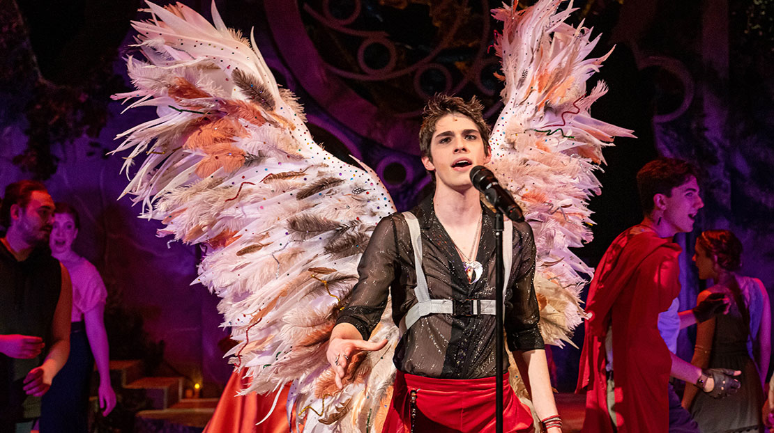 An actor sings in front of a microphone on stage dressed in red and black and wearing a large pair of feathered wings.