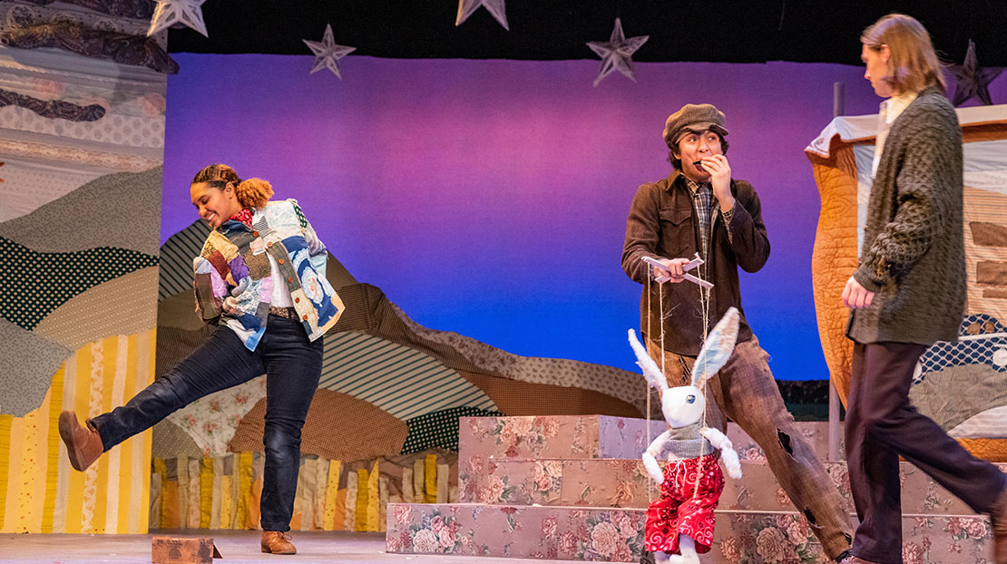 Three actors are on a stage decorated with quilt designs. One is dancing and one is playing a harmonica while holding a rabbit marionette.