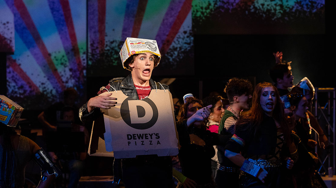 n actor, center stage, sings surrounded by an ensemble. He is dressed in a cardboard pizza box hanging over his clothes and a cardboard hat. The back of the stage has a large starburst design on it.