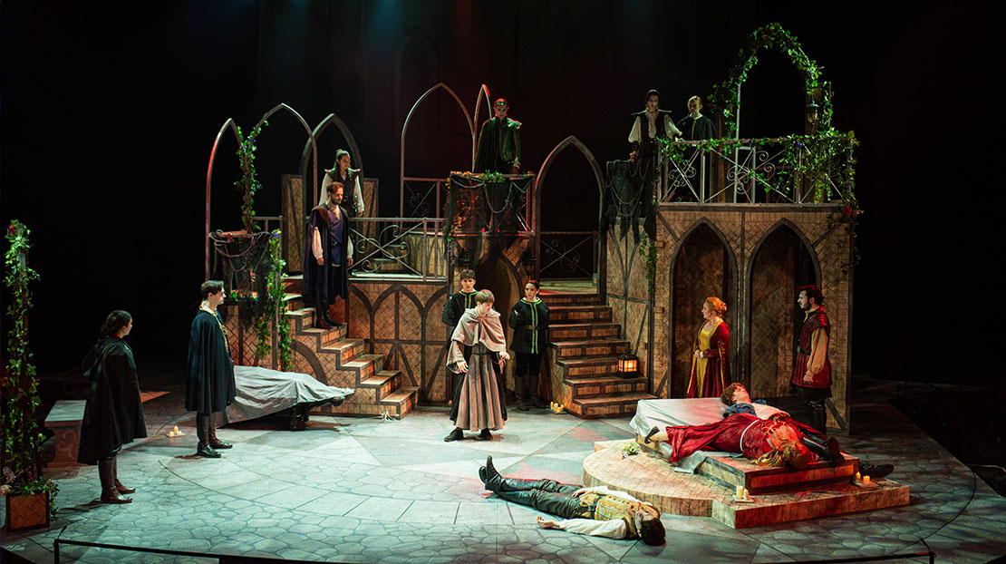 The stage is set with multiple, multi-level staircases and archways. Actors surround the center of the stage which has a bed. Two people lay dead on the bed and another on the floor beside them.