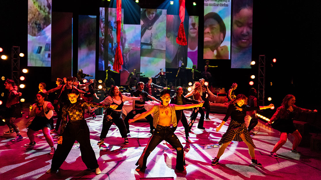 A large ensemble cast sings with their arms and legs out. The stage lighting is moody red and yellow. There are large, rectangle panels hanging from the ceiling in the background and different faces are projected onto each panel.