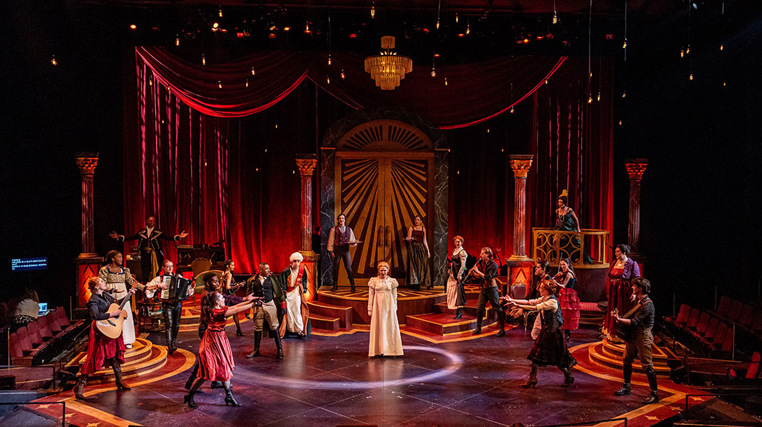 A large ensemble of actors singing, dancing, and playing instruments surround a woman in a long white dress in the center. The stage has a large door with red velvet curtains on the side and four pillars in the background and a glass chandelier and small floating lights hang from the ceiling.