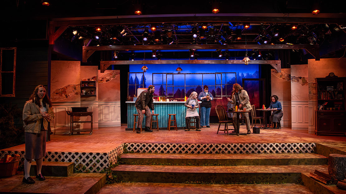 Stage is set like a rustic restaurant with a bar in front of a window view of a pine forest set against an evening blue sky. Six actors stand and sit in the restaurant.
