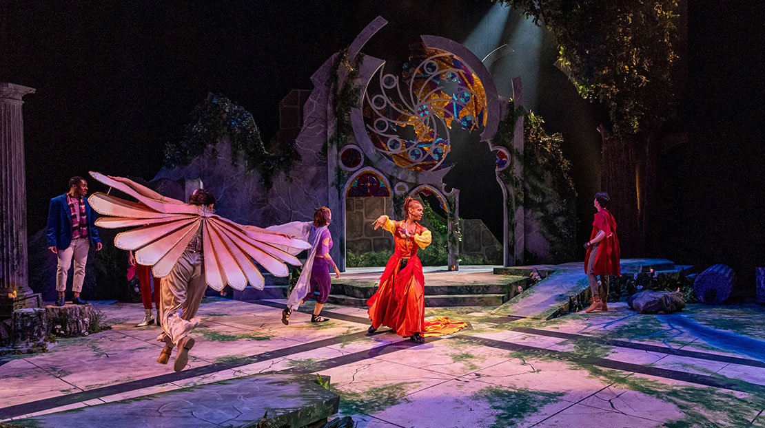 An actor in a whimsical, flowing red and yellow gown dances in the center surrounded by actors, two wearing wings. The scene displays a crumbling stone wall with large stained-glass windows and arches, as well as ivy, moss, and trees.