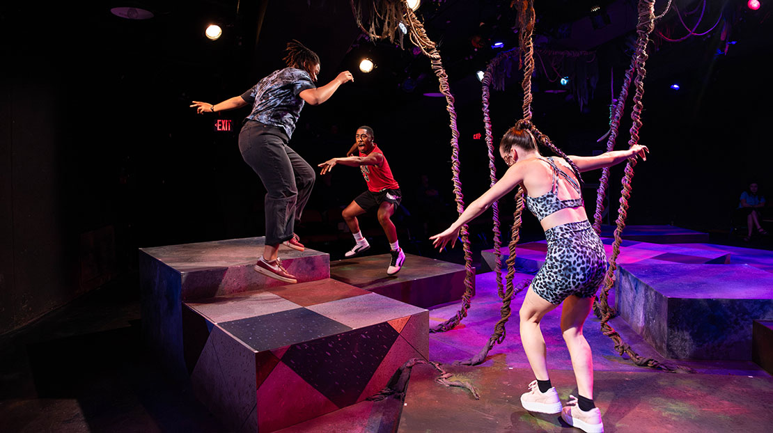 Three characters dance with their arms out around a multi-tiered stage with a tree in the middle.