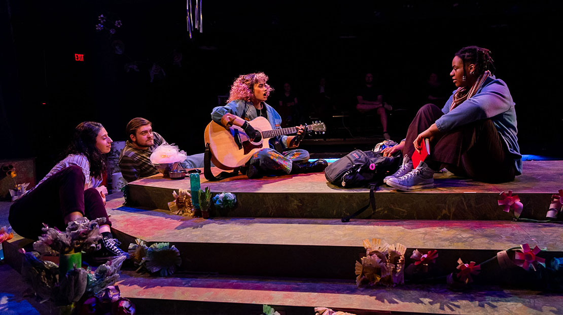 A girl sings and plays guitar on a simple tiered stage with minimal paper decorations. Three other actors sit watching her.