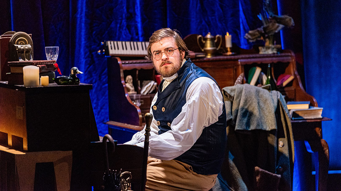 A man in period clothing plays a piano behind him hangs a blue curtain with a wooden desk in front. The desk is crammed with knick knacks on it.