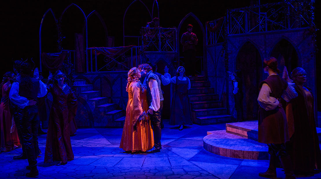 The stage is dimly lit in a dark, vibrant blue. In the center, two actors in period clothing kiss. They are surrounded by other actors doing a couple’s dance.
