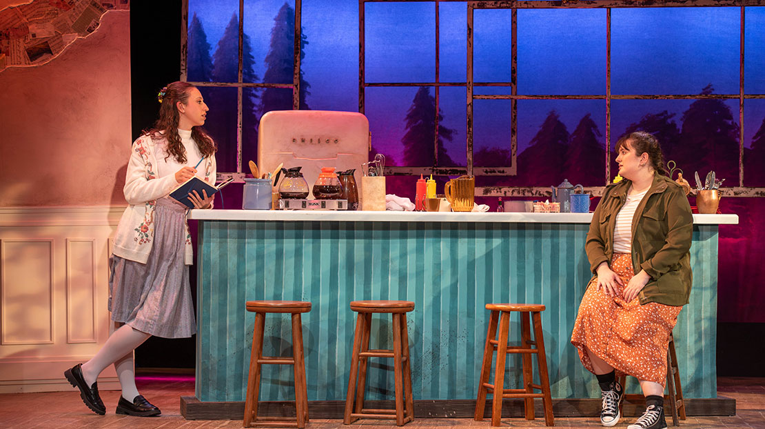  Two women are talking in a rustic bar. One is standing and she holds a book and pen. The other sits on a bar stool. The bar is blue and is in front of a winder with views of pine trees against an evening blue sky.