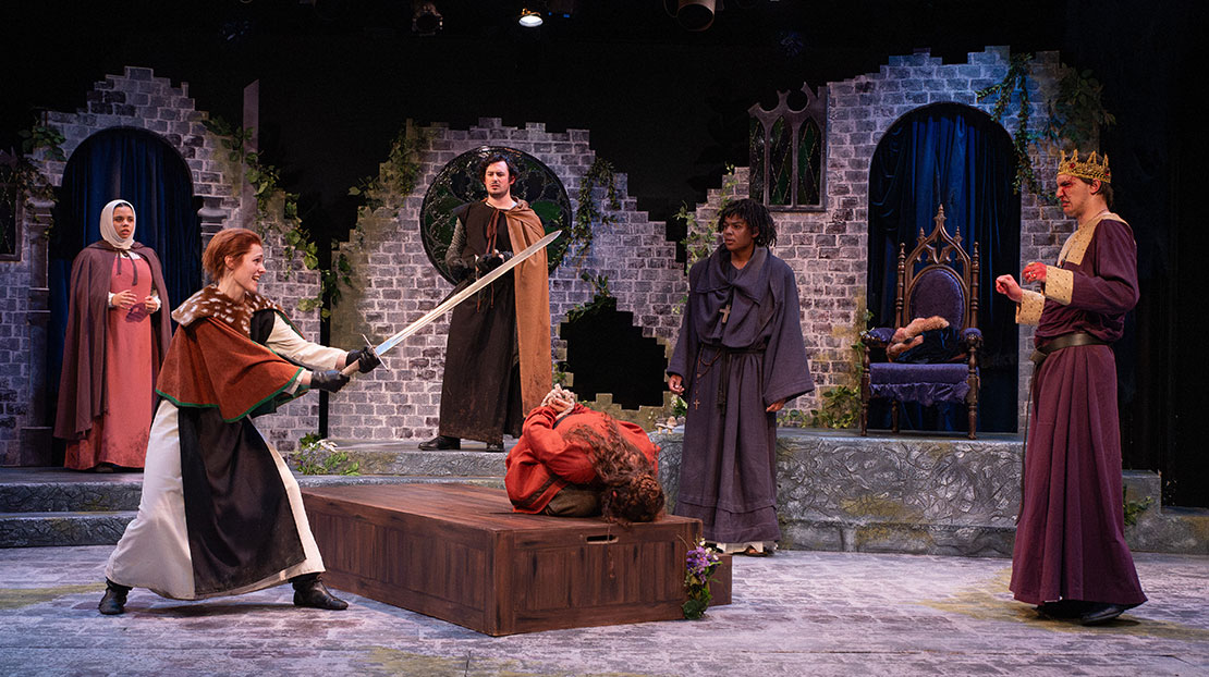 A woman is kneeling, head to the ground with her hands tied behind her back. Another actor holds a sword over her head. Four actors dressed in robes surround them watching. They are in front of a crumbling stone wall with arches and ivy. 