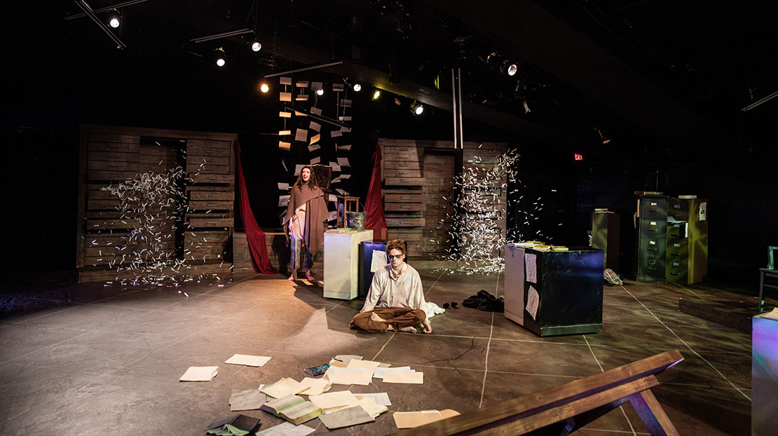 Two actors in brown and white clothing are in an office space. One sits cross-legged with his eyes closed and the other stands. Sheets of paper are everywhere, scattered all over the floor, hanging from the ceiling in the background, and paper confetti is floating on both sides.
