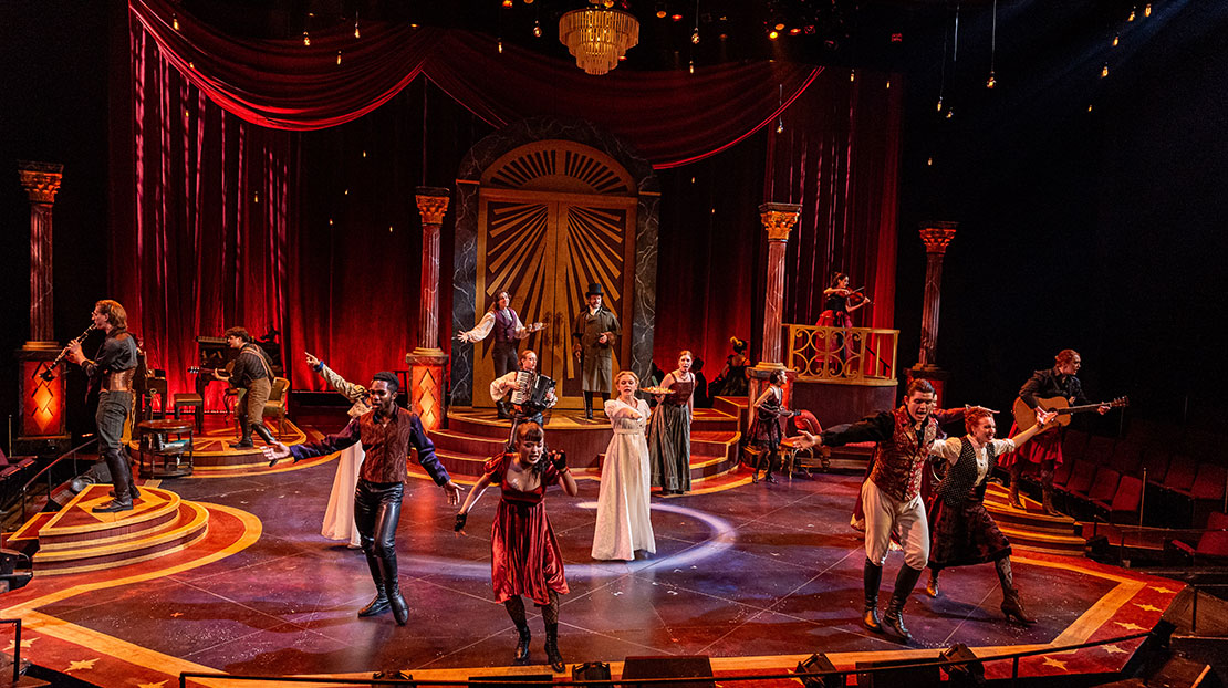 A large group of actors and musicians are singing, dancing, and playing instruments. The stage is set with low, multi-tiered. Stairs, red velvet curtains, pillars and a large door in the center.
