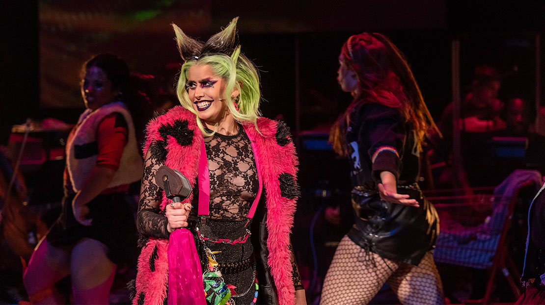 An actor smiles. She has bright green hair coming to two points on top resembling horns. She wears black lipstick and eye makeup and is dressed in a hot pink, furry vest and black lace top.