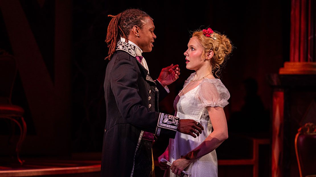 A couple face each other about to embrace. They are dressed in period clothing. His hair is pulled into a ponytail and she wears her hair in an updo pinned with flowers.