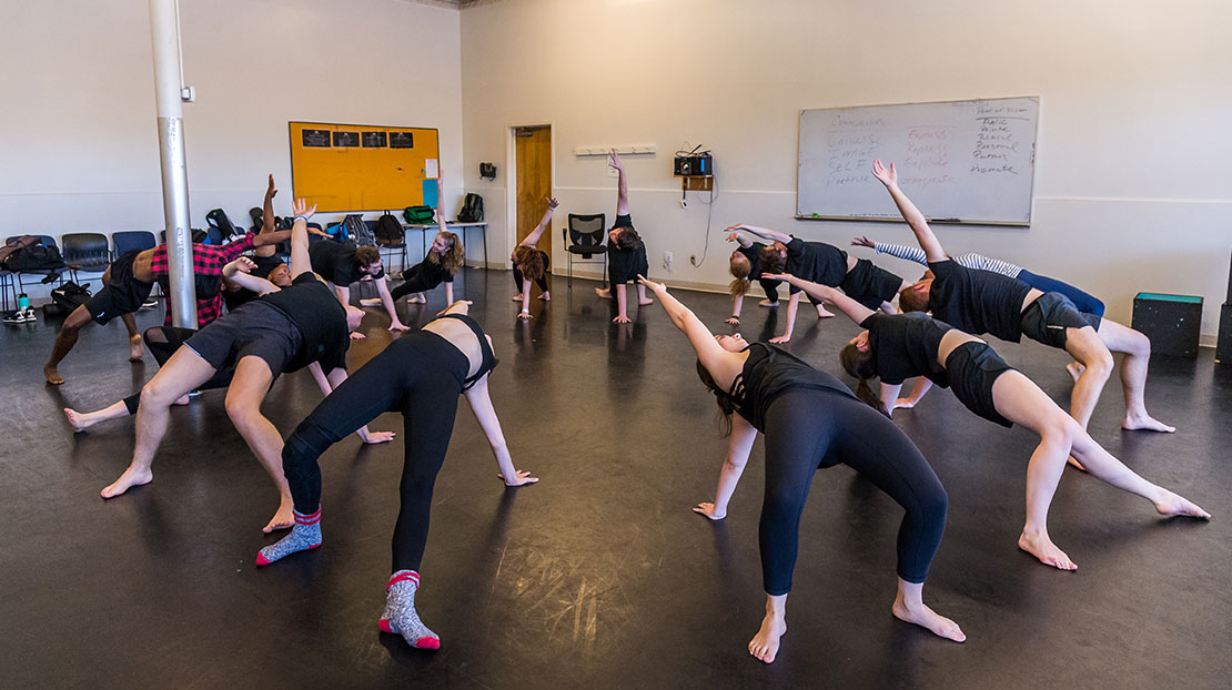 Students in an acting class, all in a circle, do a one-handed bridge pose. The classroom space is a large studio with a black dance floor.