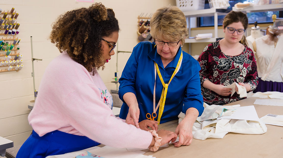 A professor guides a student with a costume construction project. They are at a worktable in a design studio, next to another student at work.