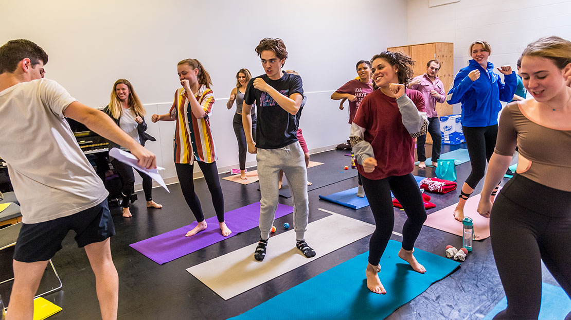 Students in a voice class are standing on yoga mats and doing a physical warm-up. 