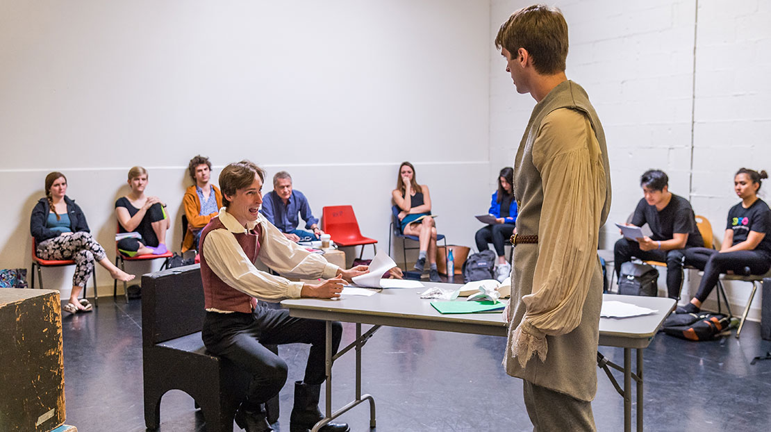 Two students wearing period clothing rehearse a scene around a table, while their teacher and classmates look on in the background.