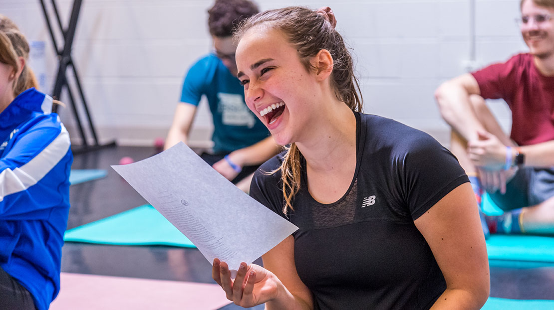 A student sitting on a mat and reading from a handout is laughing with a beaming expression.