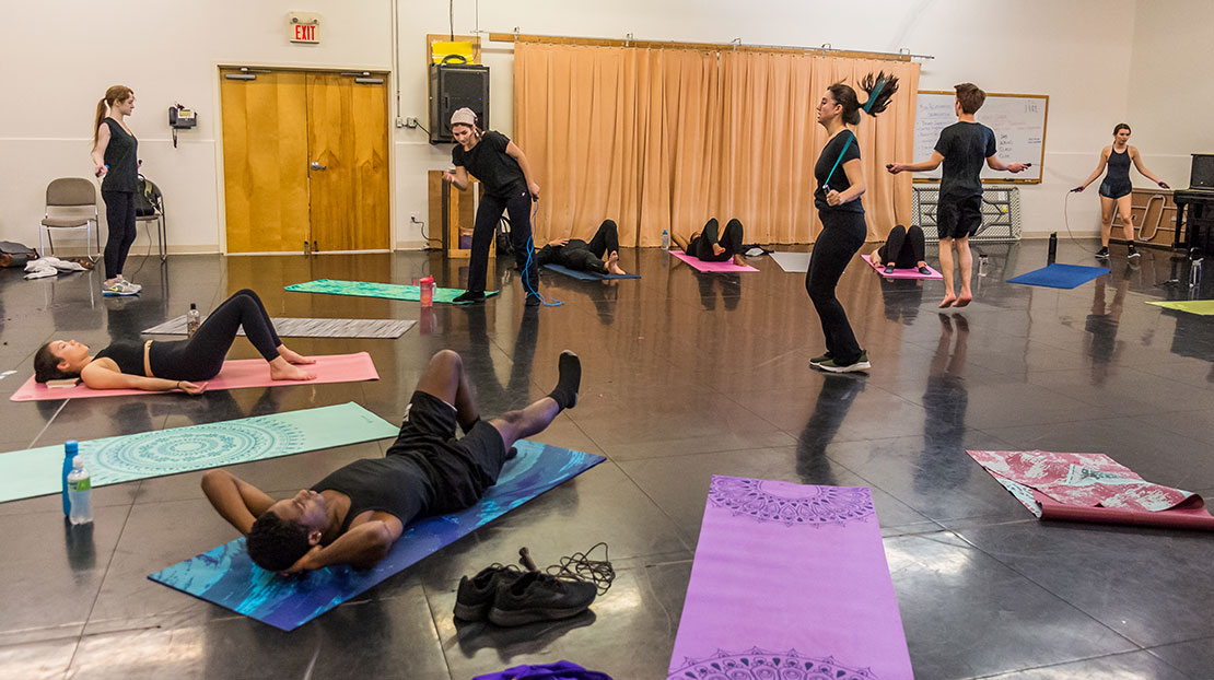 Students in a movement class, all wearing black workout outfits, do a variety of warmup activities, including jump-roping and stretching on mats. The classroom space is a large studio with a black dance floor.