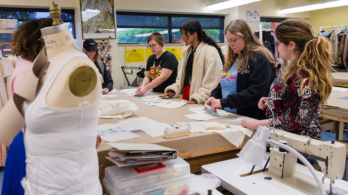 Students stand around a worktable, cutting patterns. There's a clothing form in the foreground.